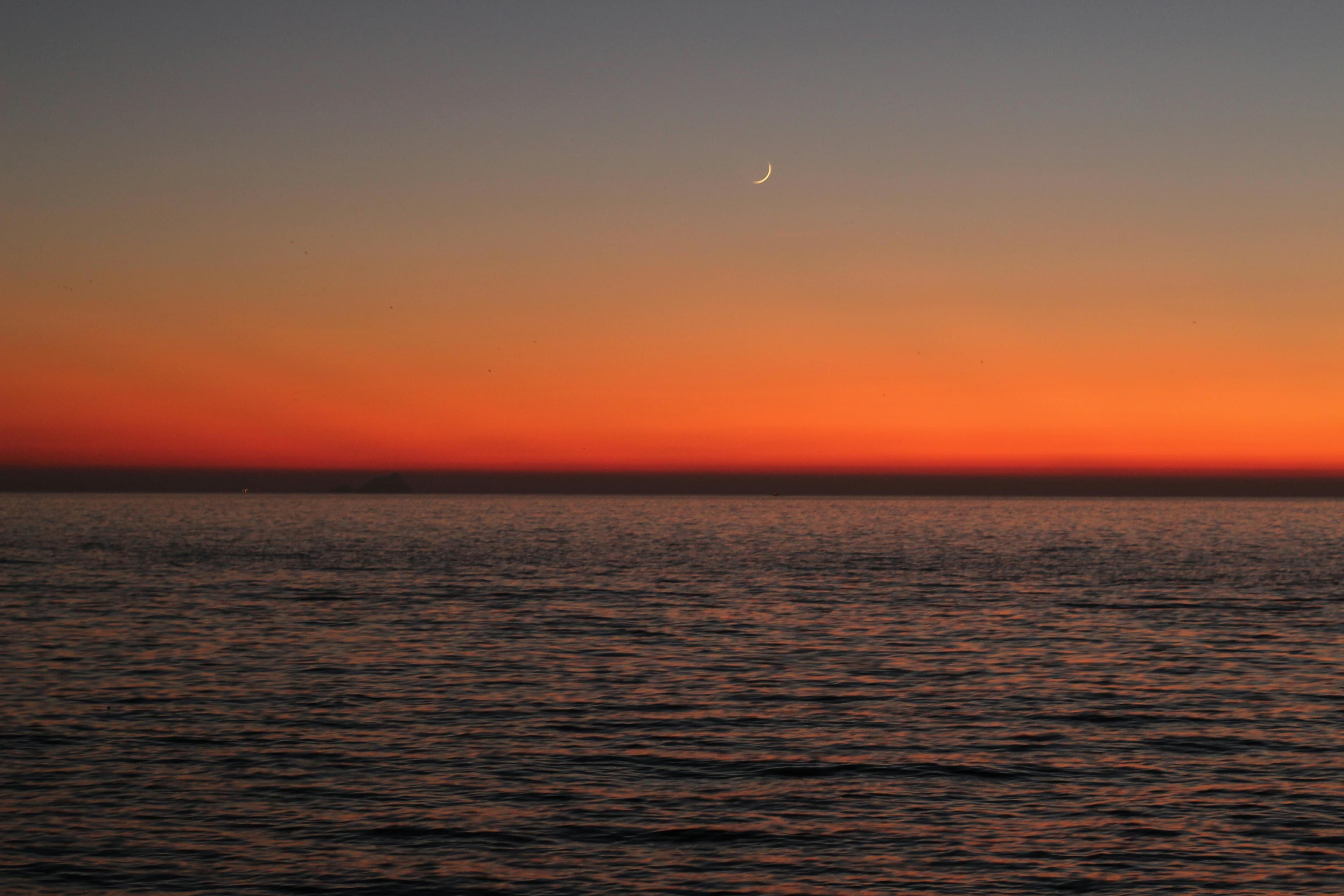 Beautiful orange sunset over calm ocean with crescent moon in sky.